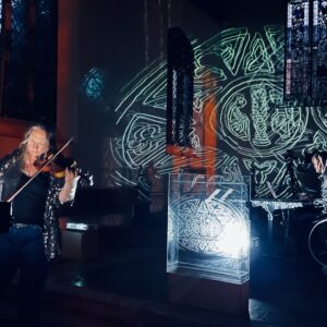 A woman plays a violin in a historic building, against a background of projected lights.